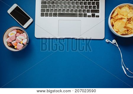 Unhealthy Work Space With Laptop, Phone, Headphones, Candy And Chips On Blue Background. Top View, S