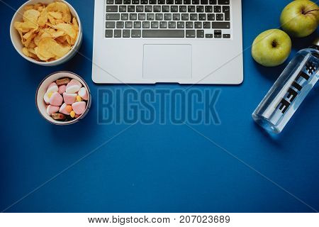 Work Space With Laptop, Candy, Chips, Water And Apple On Blue Background. Top View, Space For Text