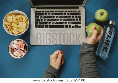 Work Space With Laptop, Candy, Chips, Water And Apple And Woman Hands On Blue Background. Top View