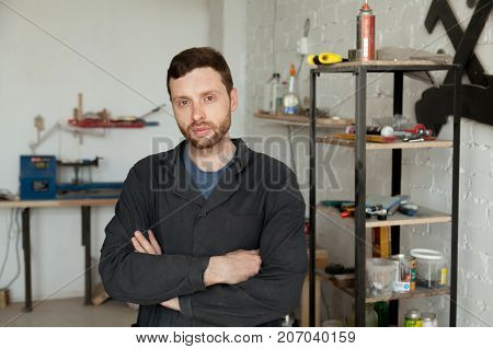 Head shot portrait of serious young successful craftsman or carpenter standing in own workshop, arms crossed looking at camera, handyman starting small carpentry workshop business concept, repair shop