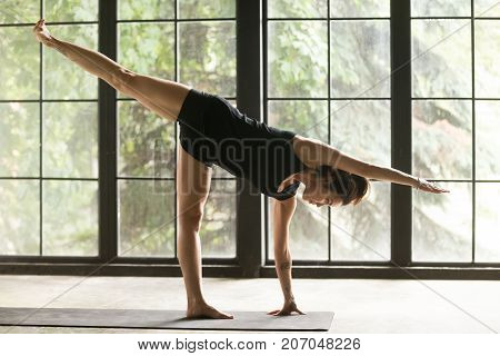Young attractive woman practicing yoga, standing in Half Moon exercise, Ardha Chandrasana pose, working out, wearing sportswear, black shorts and top, indoor full length, studio background