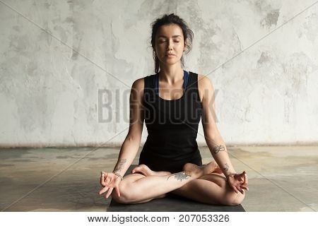 Young woman with tattoo practicing yoga, sitting in Padmasana exercise, Lotus pose, her eyes closed, working out, wearing sportswear, black shorts and top, indoor full length, studio wall background