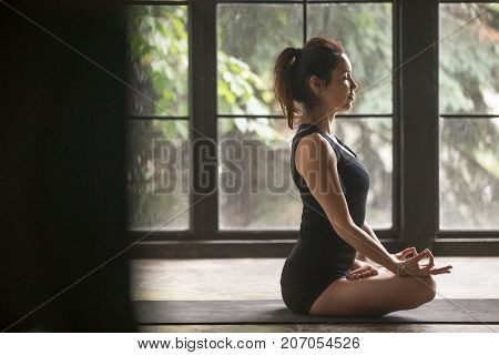 Young woman practicing yoga, sitting in Padmasana exercise, Lotus pose, mudra gesture, eyes closed, working out, wearing sportswear, black shorts, top, indoor full length, home studio life style photo