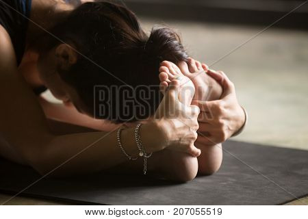 Young woman practicing yoga at home, sitting in paschimottanasana exercise, Seated forward bend pose, yogi model working out, indoor close up image, studio mat background
