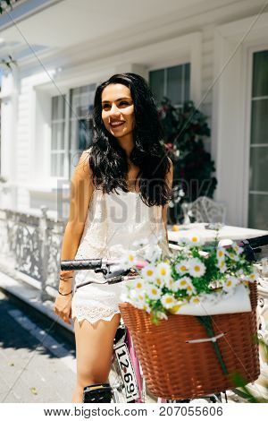 Charming woman sitting on bicycle with flowers in backet,old house on background