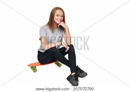 Teen girl in full length sitting on skate board smiling at camera, isolated on white background