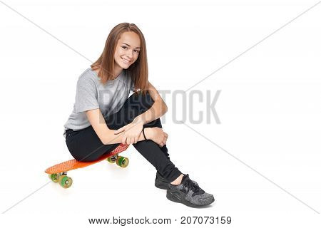 Teen girl in full length sitting on skate board smiling at camera, isolated on white background