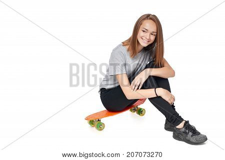 Teen girl in full length sitting on skate board smiling looking away at blank copy space, isolated on white background