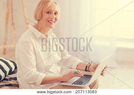 New user. Waist up of cheerful elegant senior woman sitting on her bed and using the laptop while enjoying the rest at home