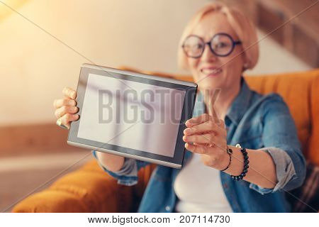Here you are. Close up of modern tablet in hands of a cheerful aged woman holding it while resting at home