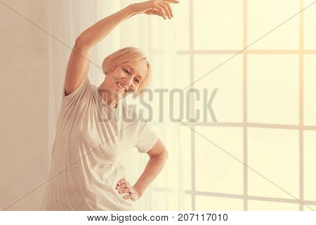 Do it with pleasure. Joyful smiling aged woman enjoying morning exercises while going in for sport at home
