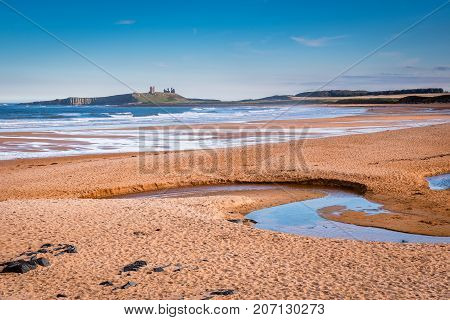 Embleton Bay and Burn, which is a wide sandy beach with the ruins of Dunstanburgh Castle providing a stunning backdrop, located on the Northumberland coastline with Embleton Burn in the foreground