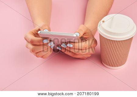 Smartphone in female manicured hands. Hands with manicure holding mobile phone, cardboard cup of coffee, pink background.
