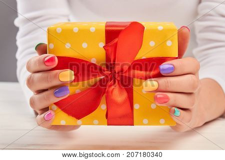 Summer manicure and yellow gift box. Woman hands with multicolored varnish on nail holding bright box with gift close up. Holidays and celebrations concept.