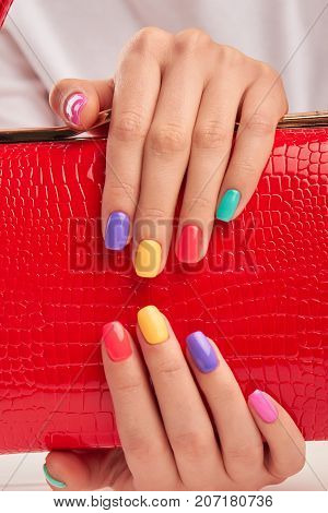 Multicolored nails and red leather handbag. Hands with beautiful summer manicure holding red lacquered handbag close up. Woman beauty and style.