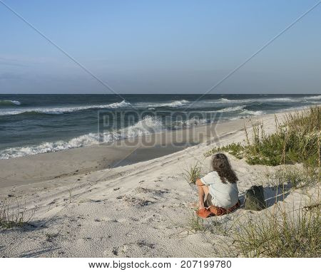 Backview of woman seated on sand dunes at Florida beach gazing out to sea at waves and crashing surf.