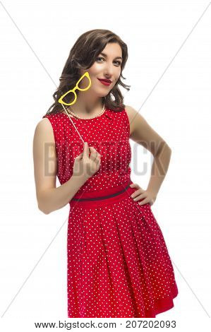 Youth Lifestyle Concept. Portrait of Sensual Alluring Caucasian Female Holding Yellow Artistic Spectacles In Front of Cheek. Posing In Red Polka Dot Dress Against Pure White.Vertical Image Composition