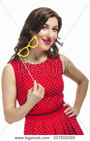 Smiling Sensual Alluring Caucasian Female Holding Yellow Artistic Spectacles In Front of Cheek. Posing In Red Polka Dot Dress Against Pure White.Vertical Image Composition