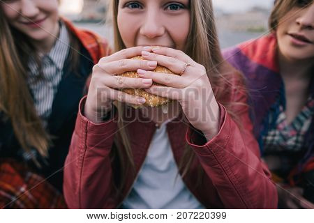 Young hot-eyed girl eats burger, close up. Meeting friends with takeaway junk food. Overnutrition and diet failure concept, mood swing in eating