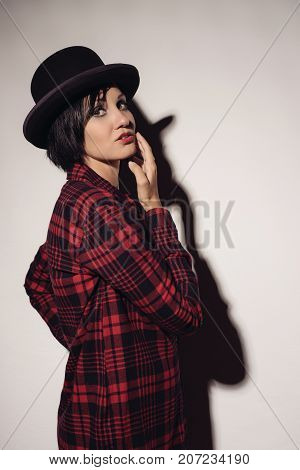 Attractive girl in red checkered shirt and hat bowler stands near white wall raising her hand to face