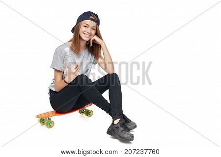 Teen girl in full length sitting on skate board holding a bottle with water smiling at camera, isolated on white background