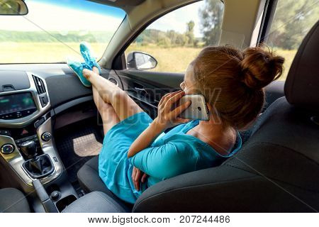 Girl in car sitting on passenger seat with feet on car dashboard and talking on mobile cell phone. Young woman relaxing with feet on dashboard. Freedom travel concept. Spending weekend in roadtrip