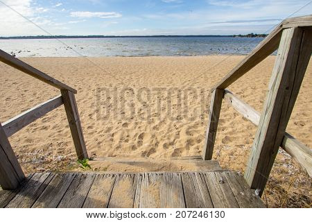 Stairs To The Beach Background. Wooden stairs lead to a wide sandy beach with a sunny blue water horizon. Color image in horizontal orientation with copy space.