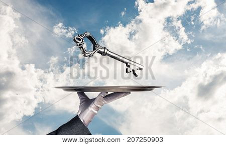 Cropped image of waitress's hand in white glove presenting big key on metal tray with cloudy skyscape on background.