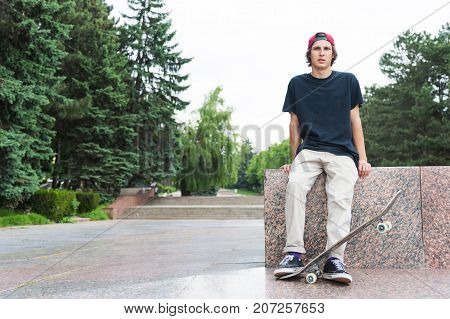 The long-haired skater in a cap and T-shirt sits with his back to the camera and thinks next to the skateboard against the background of the landscape landscape.