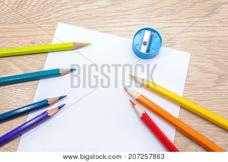 Different colored pencils paper sheet and pencil sharpener lie on wooden table. Photo with space for text. Seven pencils of rainbow colors. Copyspace. Back to school. Wooden background.