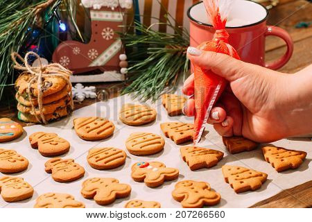 Icing process of Christmas bakery. Unrecognizable woman decorates homemade gingerbread cookies on cooking sheet, near pine branch with festive decor. Family culinary and New Year traditions concept