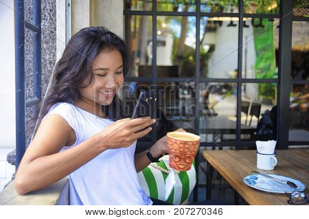 young beautiful and happy Asian woman smiling relaxed enjoying breakfast sitting outdoors at coffee shop holding mug and mobile phone networking having social media internet fun