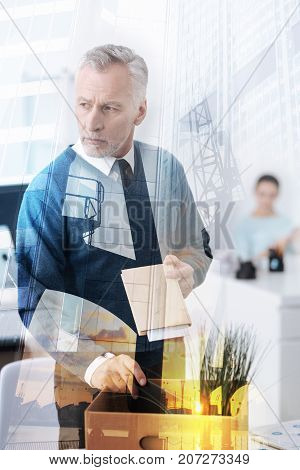 Time to leave. Serious morose adult man standing near table and packing his belongings while leaving this job position