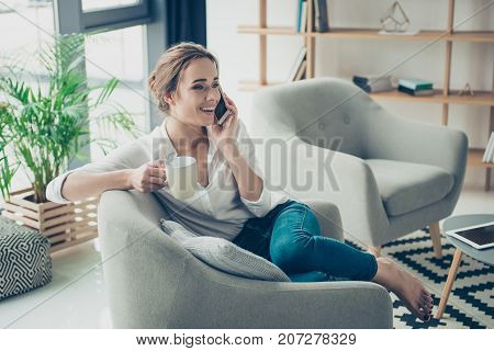 Comfort and cozy girlish time. Cute lady in smart trendy wear is chatting on her telephone sitting on armchair. Smiling sitting in relaxing atmosphere indoors at home with plants and nice design