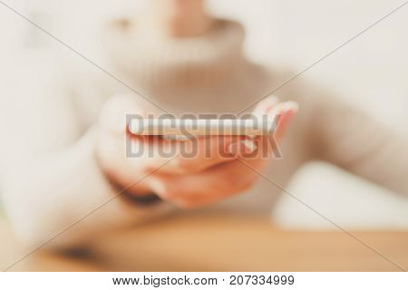 Woman holding smartphone while sitting at wooden table, selective focus, close up.