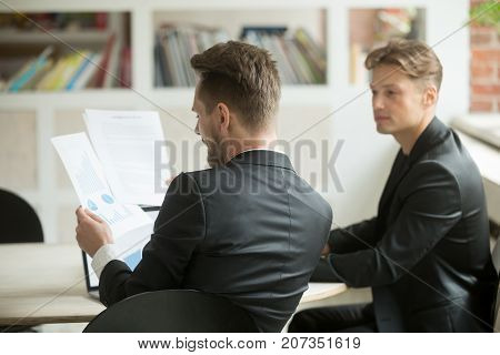 Two young businessmen coworkers analyze financial stats report at desk in modern office. Project manager and CEO overseeing corporate progress. Stock investment profit stats. Finance success concept.