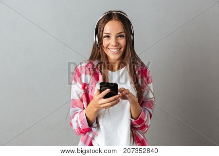 Young cheerful beautiful brunette woman with long hair, listening music with white heaphones, looking at camera, over gray background