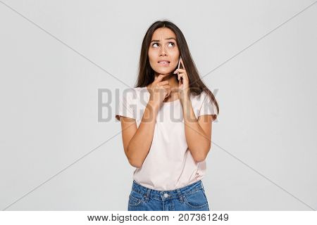 Portrait of a thoughtful doubtful asian woman talking on mobile phone and looking up isolated over white background