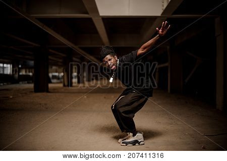 Emotional full length portrait of the dancing afro american man in the half squat with dreadlocks and dressed in black clothes