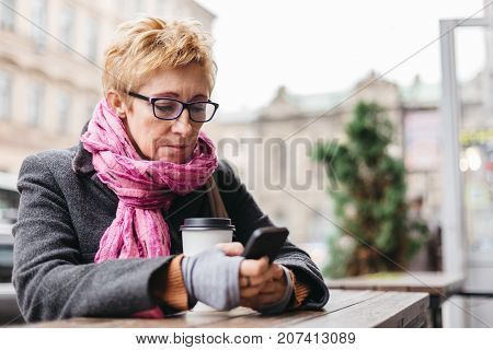 Adult blonde woman sitting in outside cafe with paper cup and surfing smartphone.