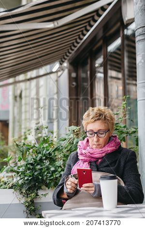 Smiling adult woman browsing smartphone and sitting with paper cup in outside cafe.