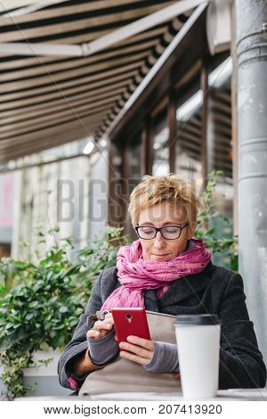 Smiling adult woman browsing smartphone and sitting with paper cup in outside cafe.