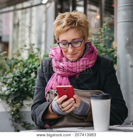 Smiling adult woman browsing smartphone and sitting with paper cup in outside cafe.