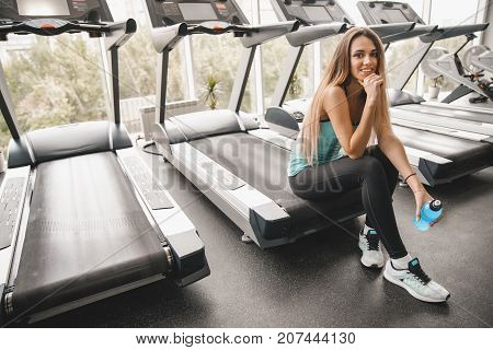Beautiful athletic girl sits on a treadmill with a bottle of energy sports drink and smiles at the camera. Sports lifestyle.