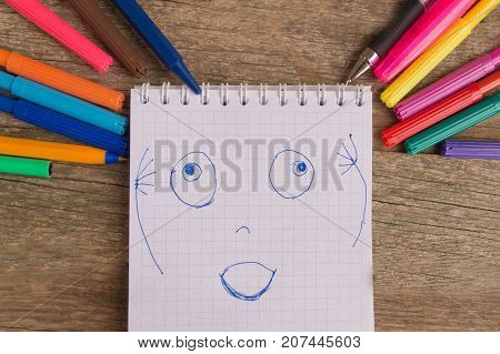 Open white notepad with colorful felt-tip pens and ball pens with drawing glad face on the wooden table.