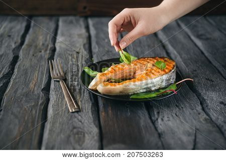 Grilled Steak Of Salmon Fish On A Black Plate. Woman Puts Leaf Of Salad On A Dish With Right Hand.
