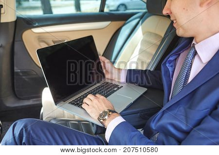 Close-up of a young businessman who uses a laptop to work in the backseat of a car.