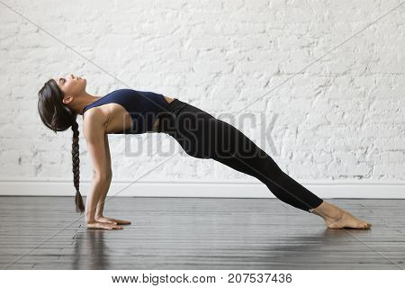 Young attractive woman practicing yoga, stretching in Purvottanasana exercise, Upward Plank pose, working out, wearing sportswear, black top and pants, indoor full length, studio background