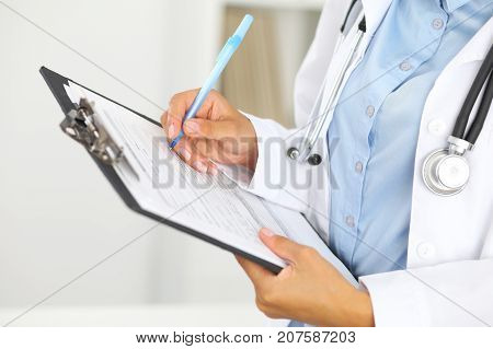 Close-up of a female doctor filling  up medical form at clipboard while standing straight in hospital.