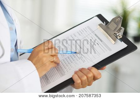 Close-up of a female doctor filling  up medical form at clipboard while standing straight in hospital.